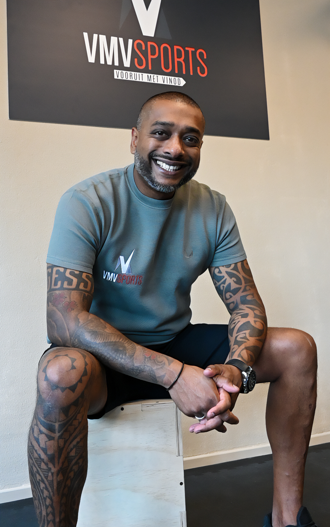 Portret van Vinod Nagessar, lachend in de VMV-studio met het VMV Sports-bord achter hem aan de wand.