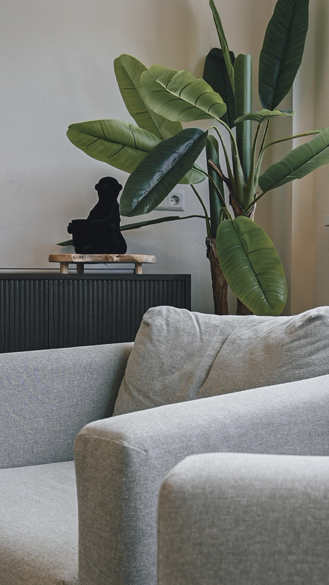 Detail van de gespreksruimte: de armleuning van een fauteuil met daarachter een plant en een beeldje op het dressoir