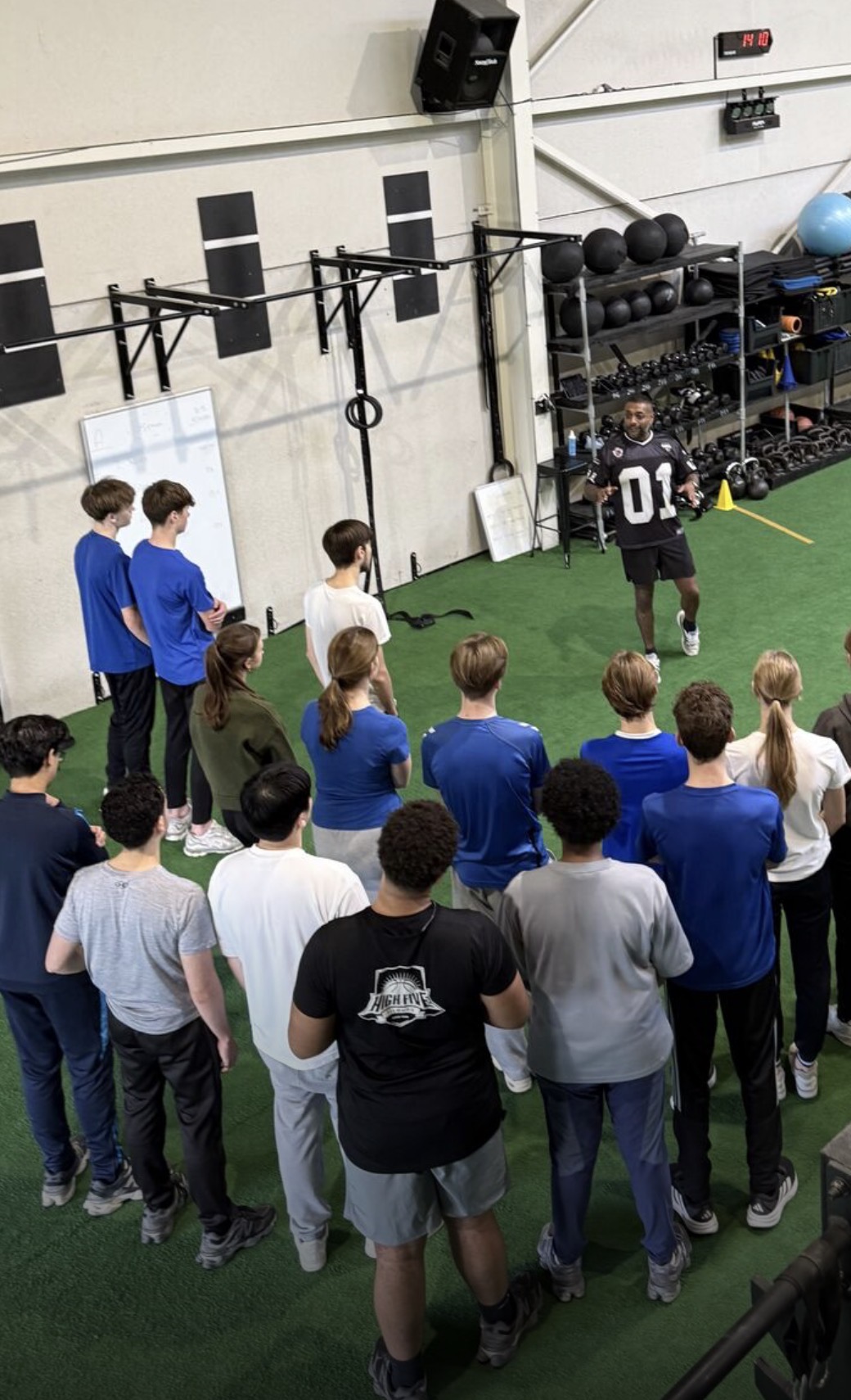 Vinod geeft training aan een grote groep leerlingen in een sporthal en legt staand voor de groep iets uit.