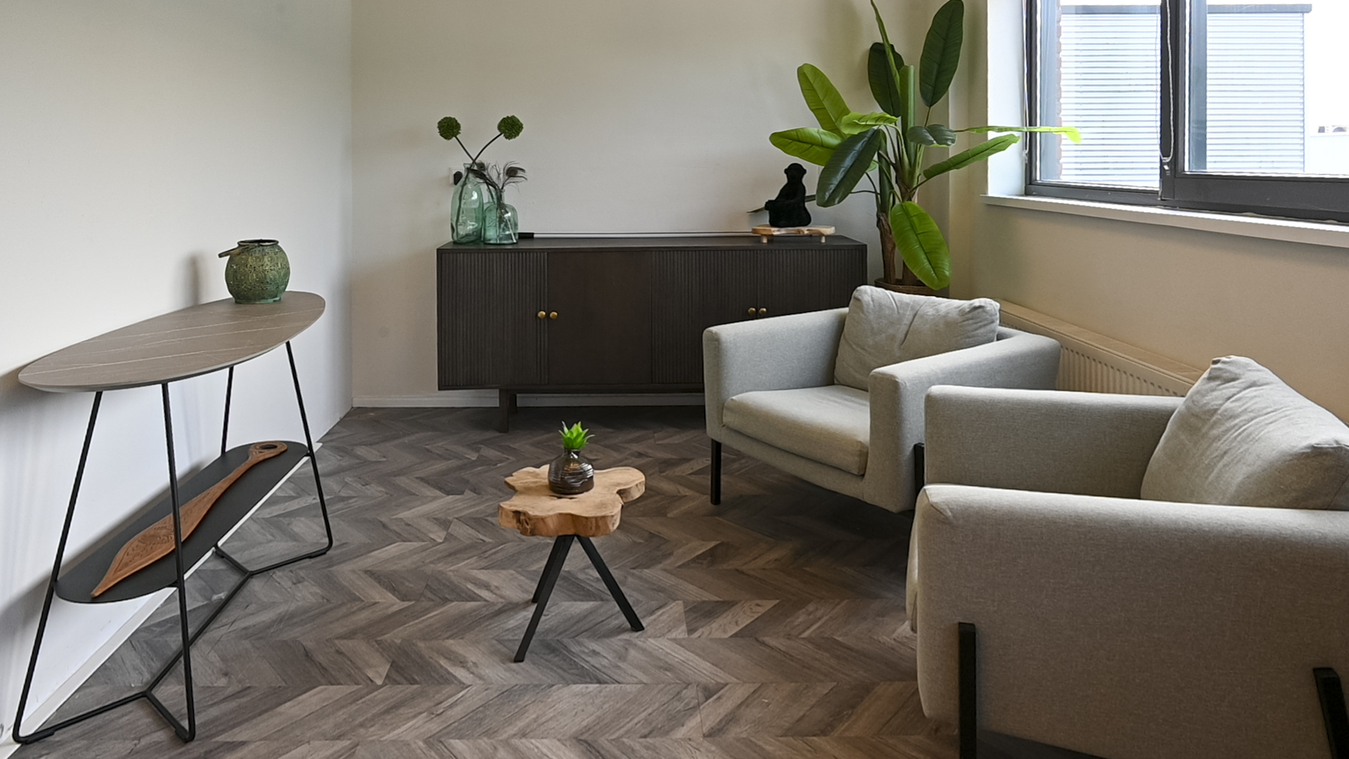 De gespreksruimte van VMV met lichte fauteuils, visgraatvloer, dressoir en een grote plant bij het raam