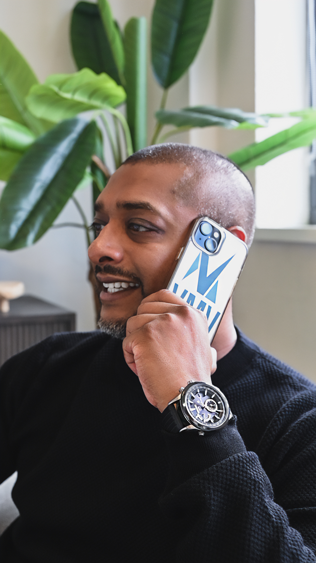 Vinod belt lachend met een telefoon met VMV-hoesje in de gespreksruimte, met groene planten op de achtergrond.